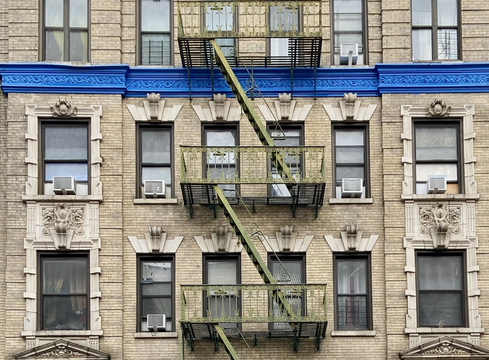 Screenshot 2023-05-18 at 3.52.32 PM.png The facade of a residential prewar brick building with a green fire escape and a bright blue cornice in West Harlem, Manhattan, New York City