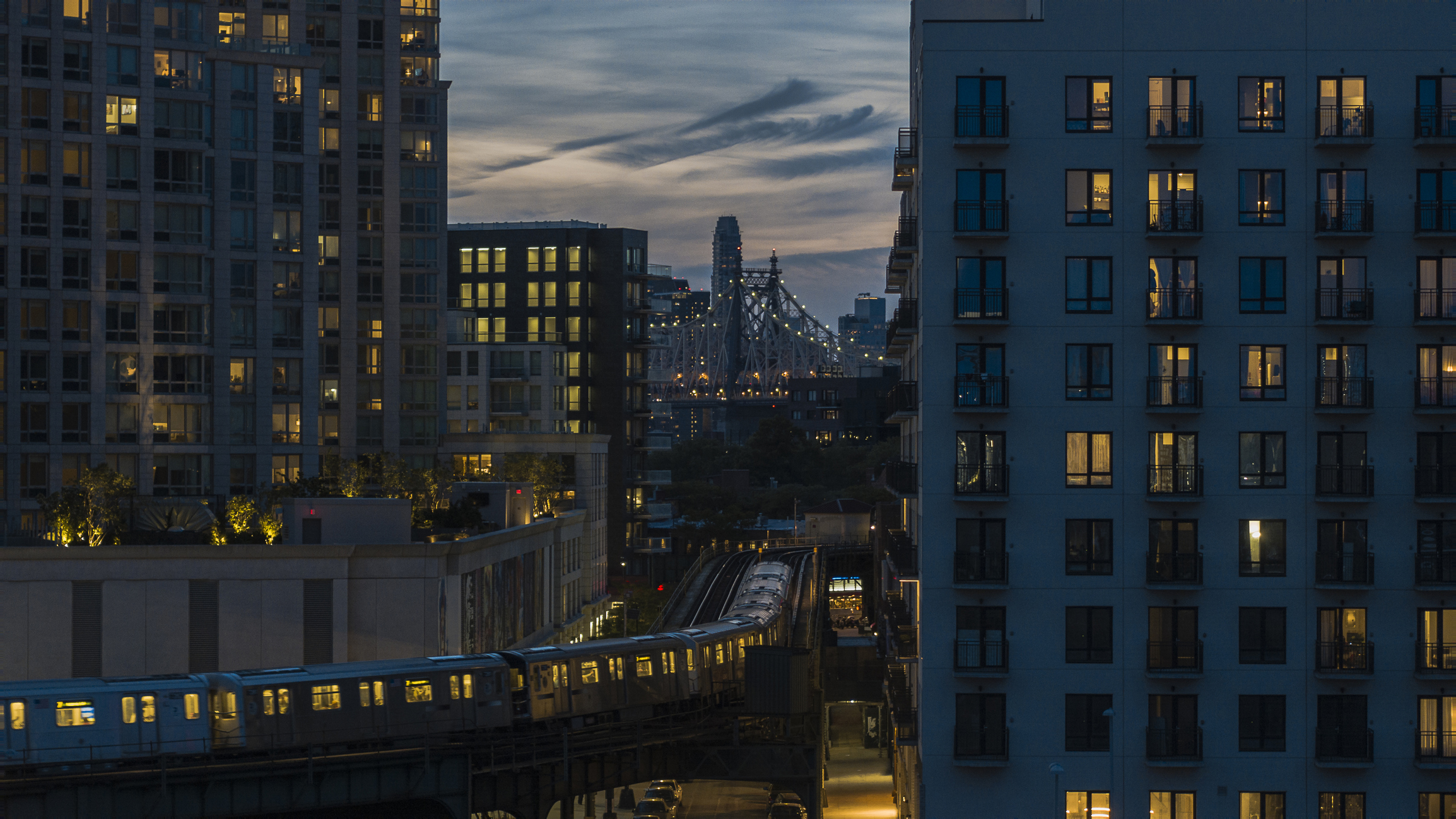 An elevated subway train riding on the line in Long Island City, Queens with Queensboro Bridge behind.