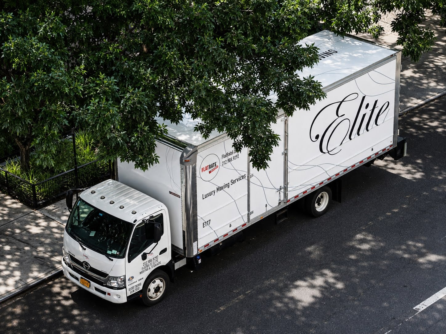 A FlatRate Elite moving truck parked on a tree-lined New York City street.