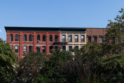 A row of colorful old brick apartment buildings surrounded by green trees during the summer in Harlem of New York City