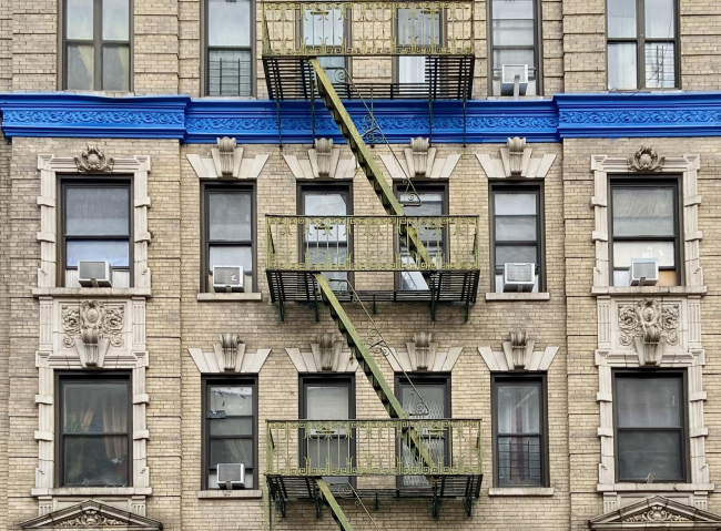 Screenshot 2023-05-18 at 3.52.32 PM.png The facade of a residential prewar brick building with a green fire escape and a bright blue cornice in West Harlem, Manhattan, New York City
