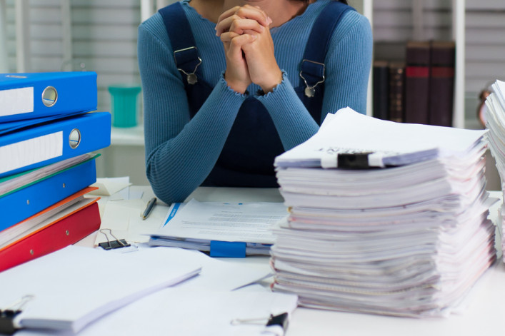 A woman clasps her hands at the center of two huge piles of paperwork.