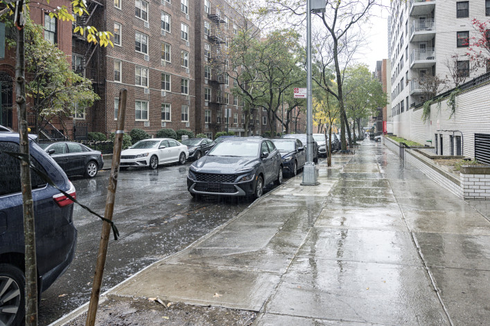 Drenched cars parked in a rain downpour in uptown New York City. Apartment buildings and brownstone row houses line this upper east side Manhattan residential district one way street and sidewalk scene.