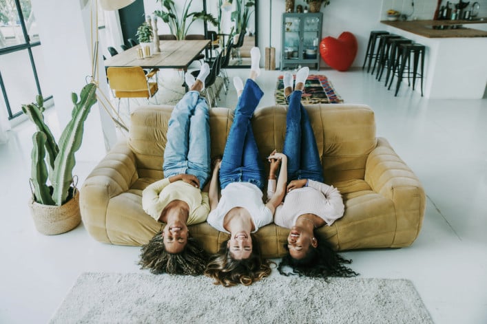 Three women lounging amusingly upside-down on a couch in a clean, brightly lit apartment