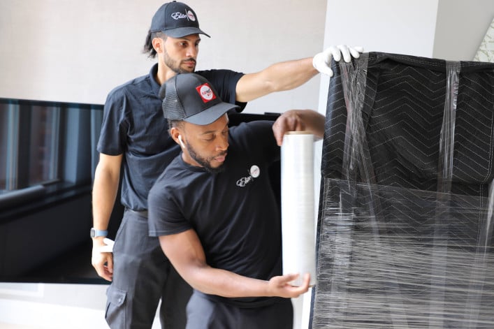 White-glove movers wrapping a large item in protective film before transport.