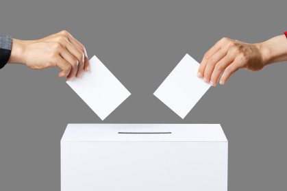 Two hands about to drop ballots into a voting box on a grey background.