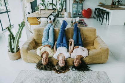 Three women lounging amusingly upside-down on a couch in a clean, brightly lit apartment