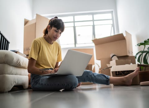 Young woman using laptop at new apartment, with moving boxes around.