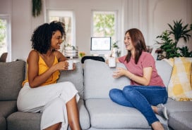 Two women sitting on sofa at home, they are drinking coffee and talking.