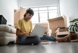 Young woman using laptop at new apartment, with moving boxes around.