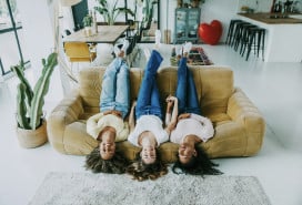 Three women lounging amusingly upside-down on a couch in a clean, brightly lit apartment