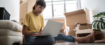 Young woman using laptop at new apartment, with moving boxes around.